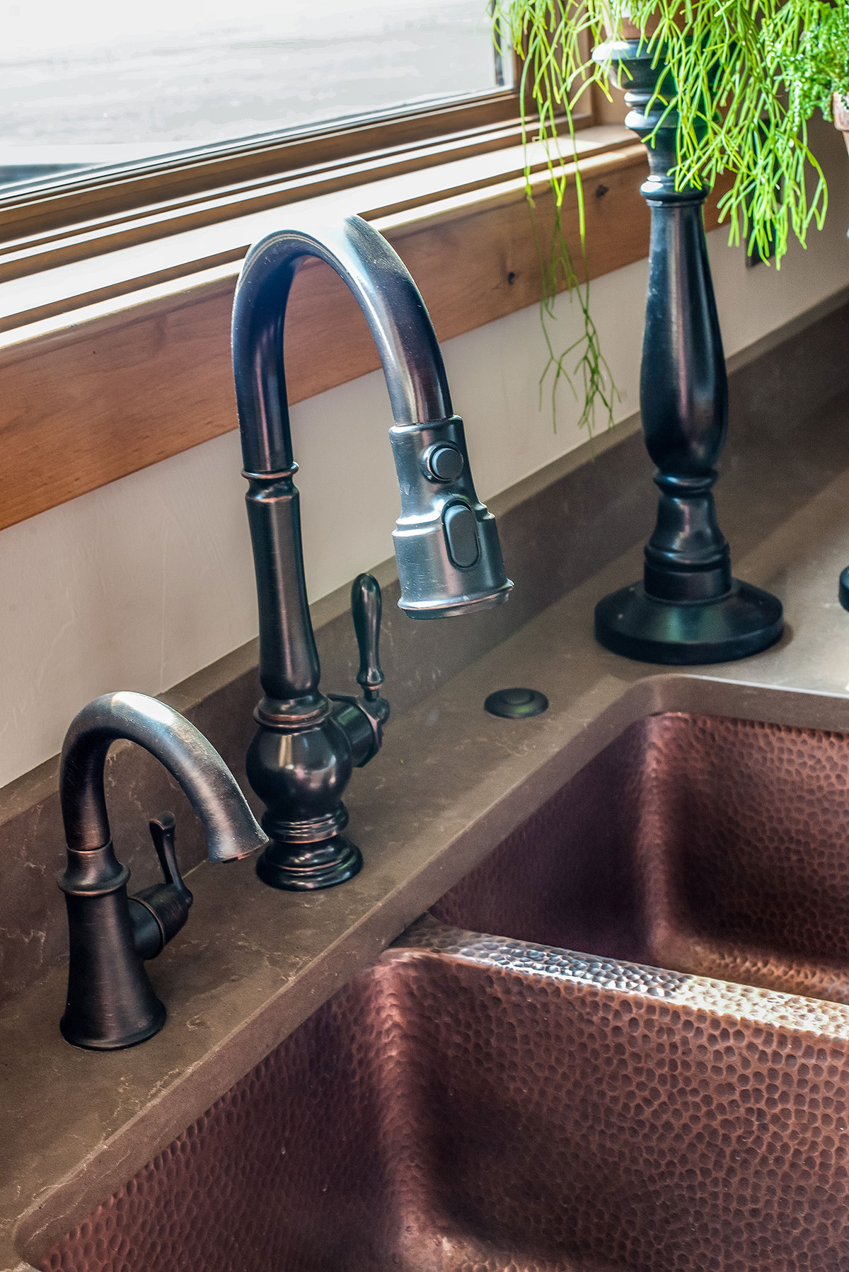 A copper double basin farmhouse sink with rubbed bronze fixtures below a window with a plant next to it