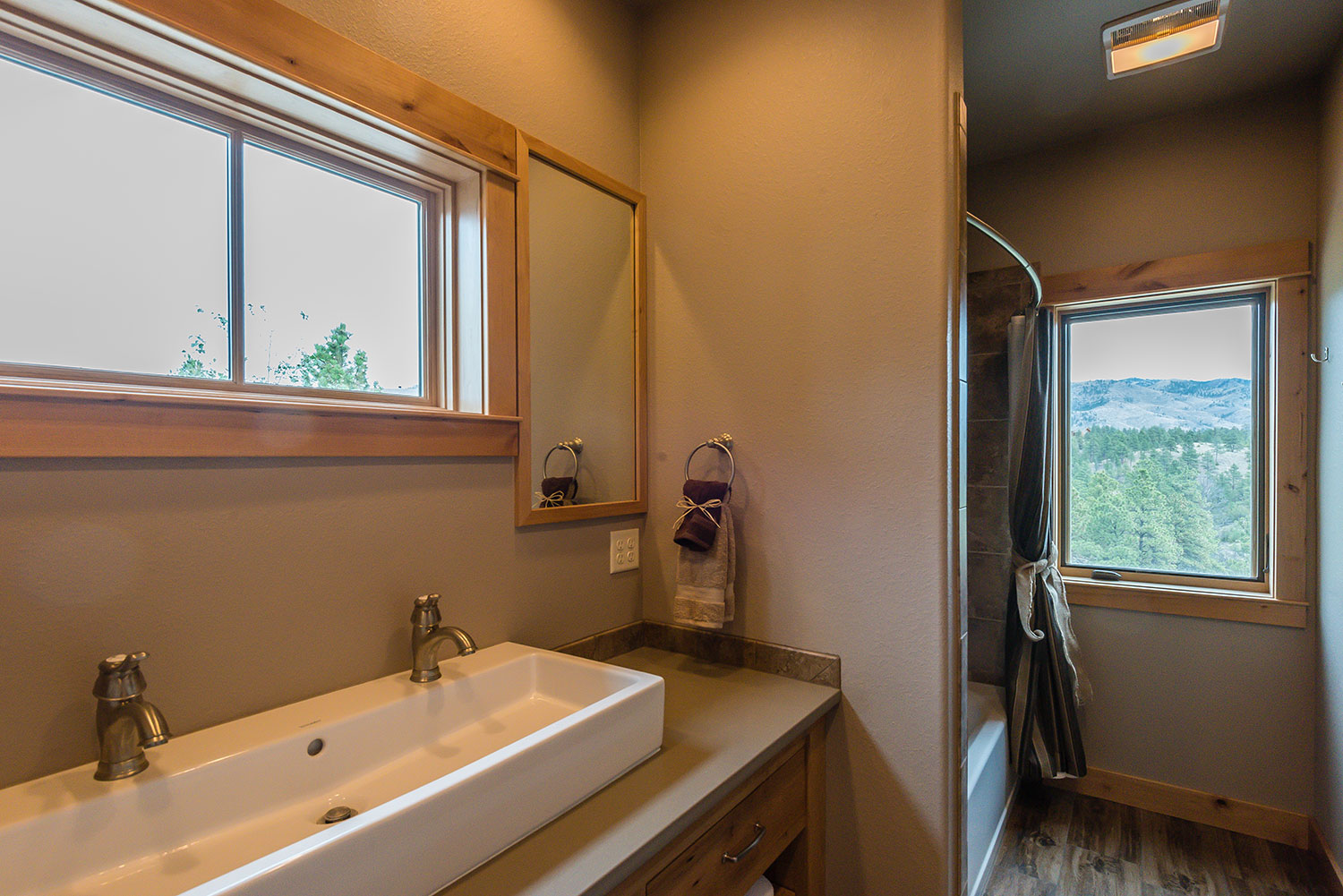 Bathroom with long rectangular sink with two faucets below a window with a shower and another window further back