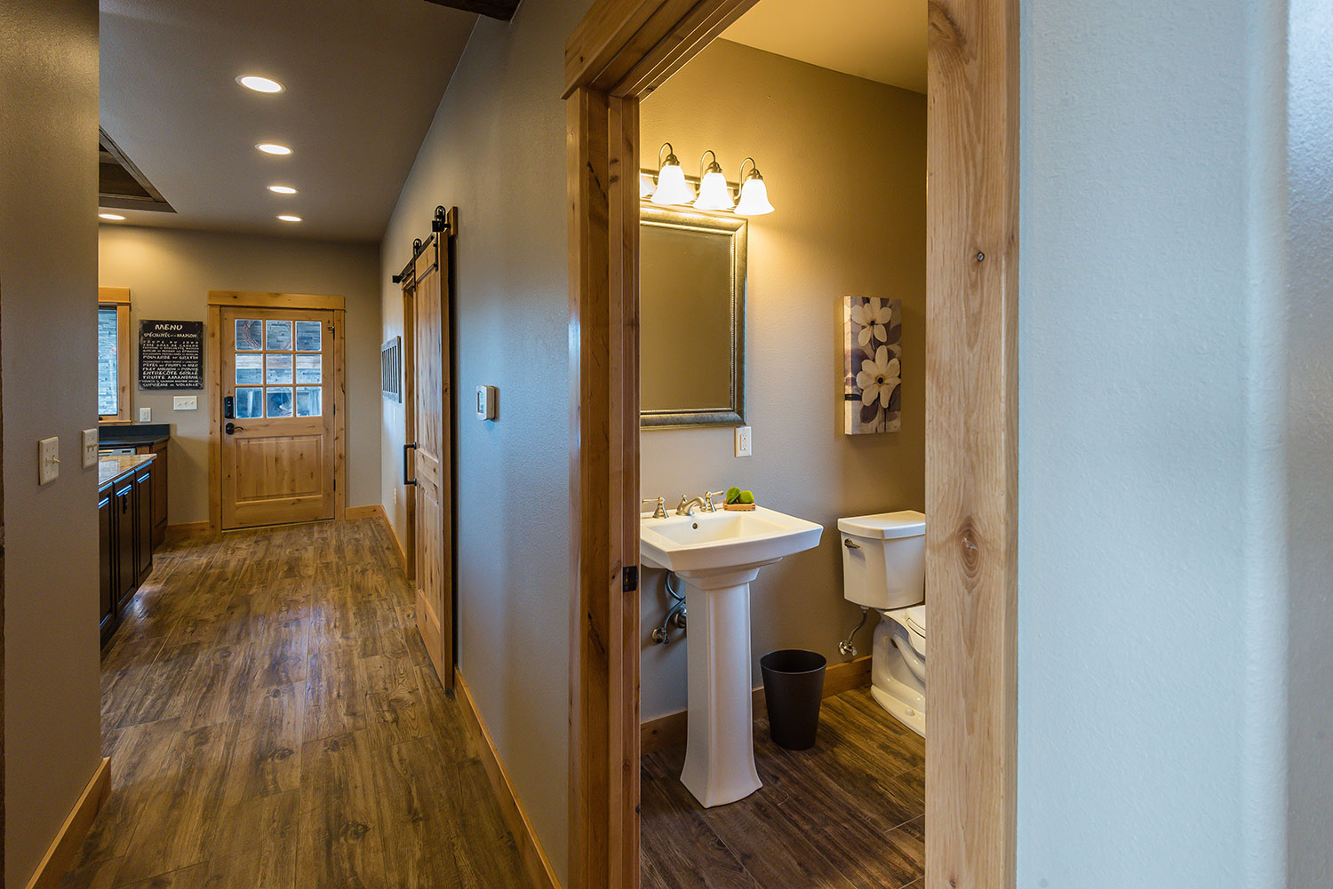 View down hallway into kitchen with a side view of a vanity with a pedestal sink and toilet in the bathroom