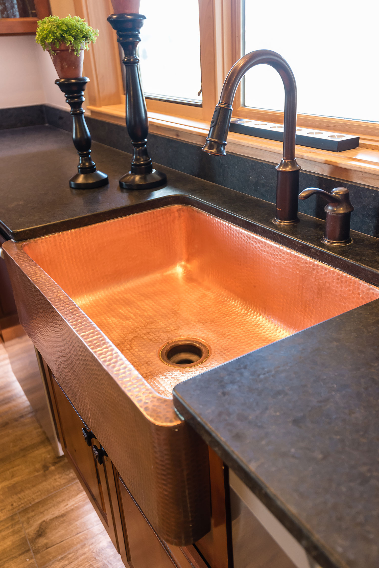 Copper farmhouse sink set in black granite countertop with rubbed bronze faucet and candles below a window