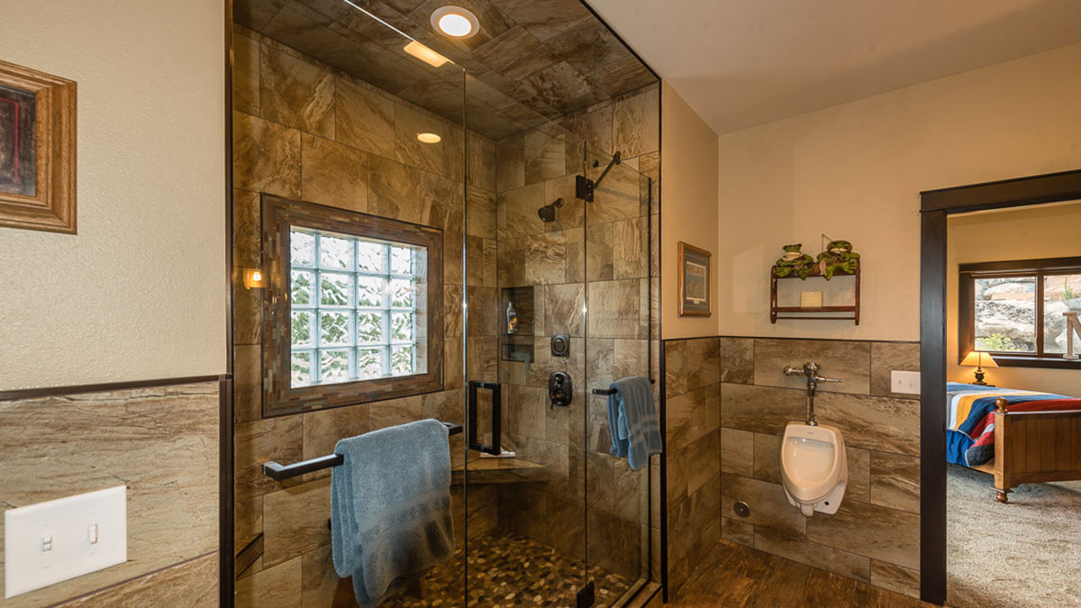 Interior of bathroom with a large walk in shower with glass door, a stand up urinal, and view into bedroom off to the left.